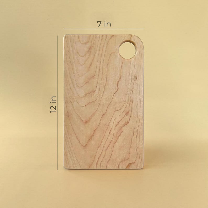 May include: A rectangular wooden cutting board with a rounded top corner and a circular hole. The board is 12 inches long and 7 inches wide. The wood grain is visible. The background is a solid light yellow.
