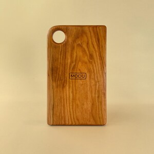May include: A rectangular wooden cutting board with a handle and a hole. The board is made of light brown wood with a visible grain pattern. The word "MODU" is printed in a small rectangle on the board.