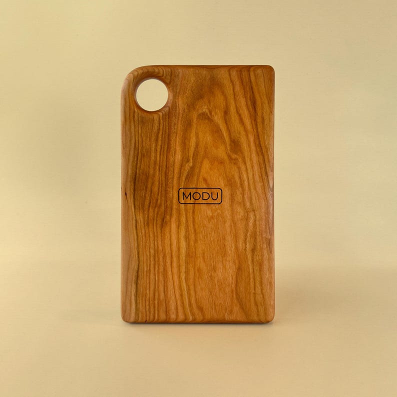 May include: A rectangular wooden cutting board with a handle and a hole. The board is made of light brown wood with a visible grain pattern. The word "MODU" is printed in a small rectangle on the board.