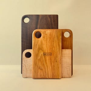May include: A collection of wooden cutting boards in various sizes and wood grains. The boards range in color from light maple to dark walnut. Each board has a rounded handle with a hole. The central board features the brand name "MODU".