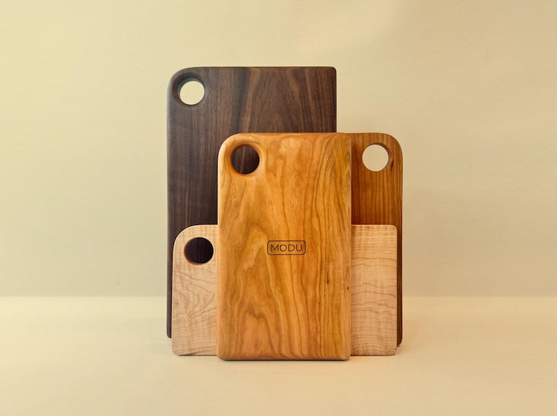 May include: A collection of wooden cutting boards in various sizes and wood grains. The boards range in color from light maple to dark walnut. Each board has a rounded handle with a hole. The central board features the brand name "MODU".
