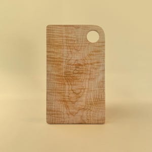 May include: A rectangular wooden cutting board with a rounded corner and a circular hole for hanging. The board has a light brown color with a unique wood grain pattern. The cutting board is approximately 12 inches long.