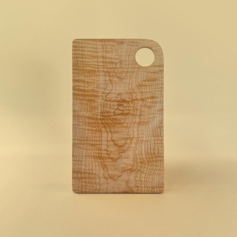 May include: A rectangular wooden cutting board with a rounded corner and a circular hole for hanging. The board has a light brown color with a unique wood grain pattern. The cutting board is approximately 12 inches long.