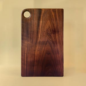 May include: A rectangular wooden cutting board with a rounded corner and a circular hole. The board is a rich brown color with a visible wood grain pattern. The word "MODU" is printed on the board.
