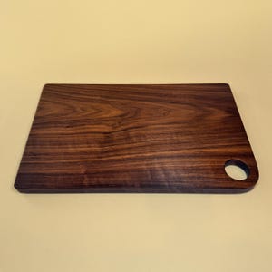 May include: A rectangular wooden cutting board with a dark brown finish. The board has a rounded corner with a circular hole for hanging. The wood grain is visible throughout the surface, adding to its natural aesthetic.
