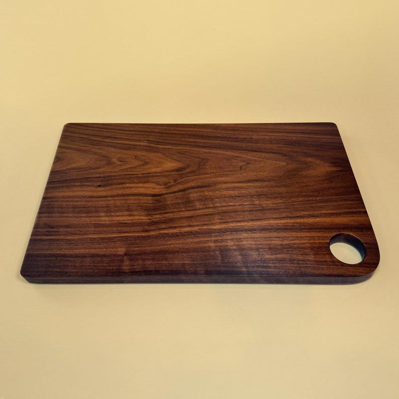 May include: A rectangular wooden cutting board with a dark brown finish. The board has a rounded corner with a circular hole for hanging. The wood grain is visible throughout the surface, adding to its natural aesthetic.