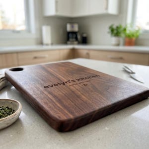 May include: A rectangular wooden cutting board with rounded corners and a handle hole. The board is engraved with the text "evelyn's kitchen made with love". The board is brown and sits on a white countertop.