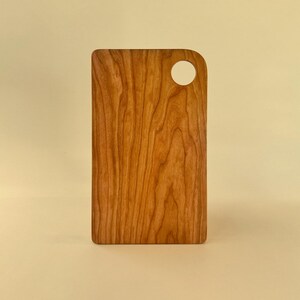 May include: A rectangular wooden cutting board with a handle featuring a circular hole. The board is made of light brown wood with a visible grain pattern. The cutting board is displayed against a light yellow background.