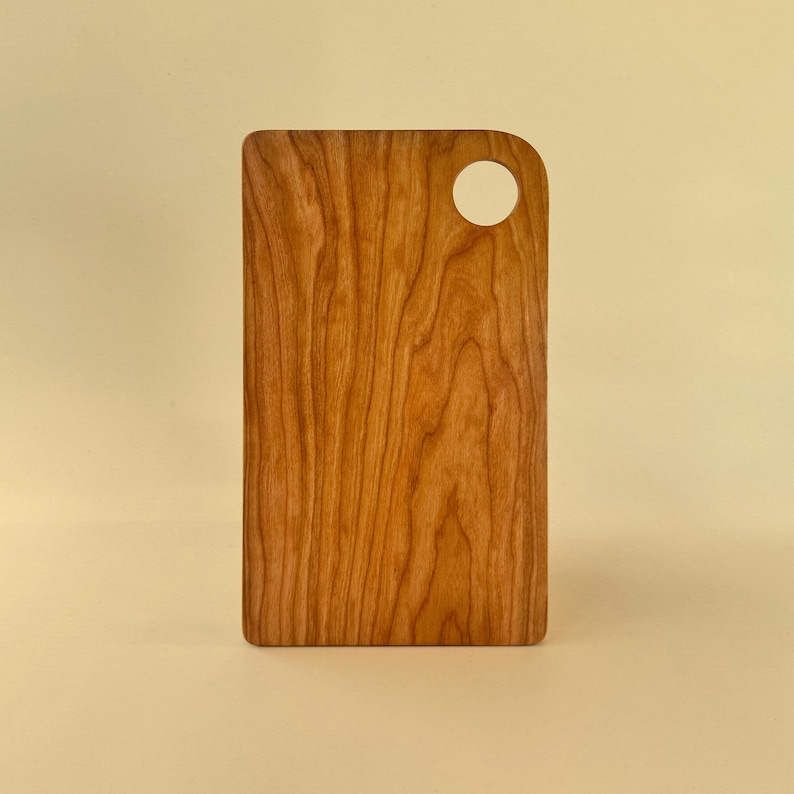 May include: A rectangular wooden cutting board with a handle featuring a circular hole. The board is made of light brown wood with a visible grain pattern. The cutting board is displayed against a light yellow background.