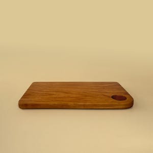 May include: A rectangular wooden cutting board with a rounded corner and a circular hole. The board is made of light brown wood with visible grain and a smooth finish. The cutting board is approximately 12 inches long and 8 inches wide.