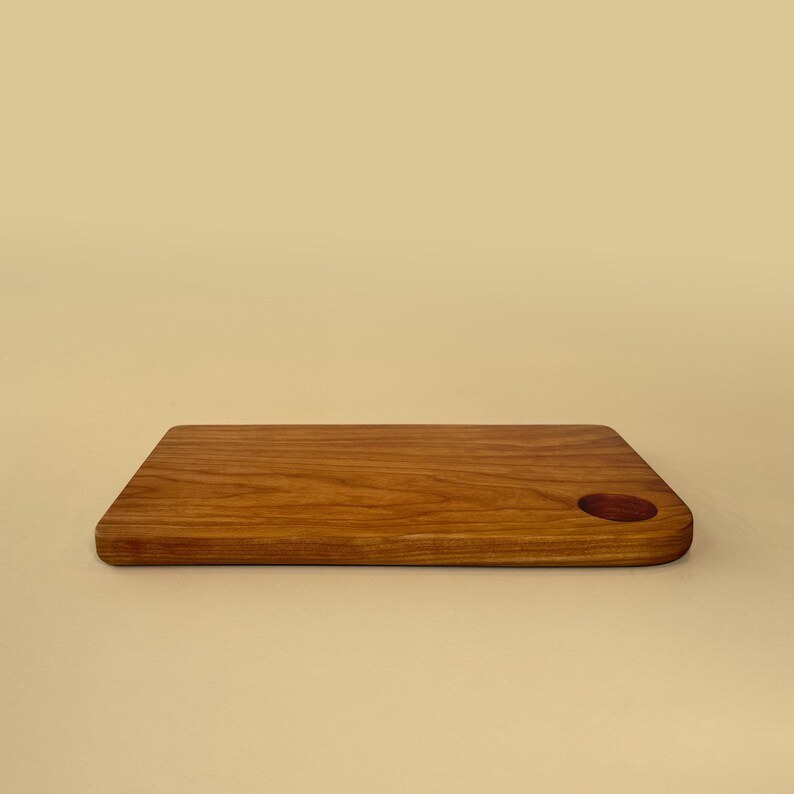 May include: A rectangular wooden cutting board with a rounded corner and a circular hole. The board is made of light brown wood with visible grain and a smooth finish. The cutting board is approximately 12 inches long and 8 inches wide.