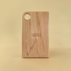 May include: A rectangular wooden cutting board with a rounded top corner and a circular hole for hanging. The board has a light wood grain pattern and the word "MODU" is printed on the front.