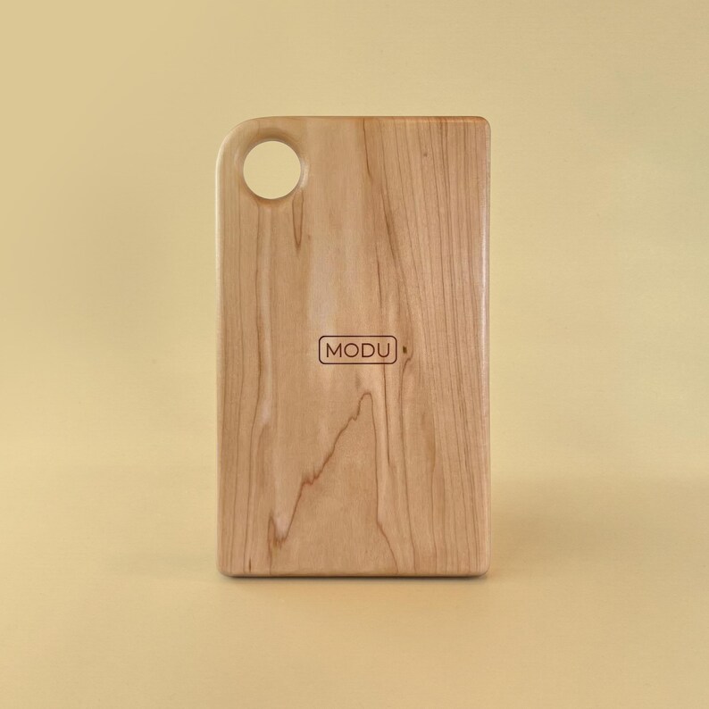 May include: A rectangular wooden cutting board with a rounded top corner and a circular hole for hanging. The board has a light wood grain pattern and the word "MODU" is printed on the front.