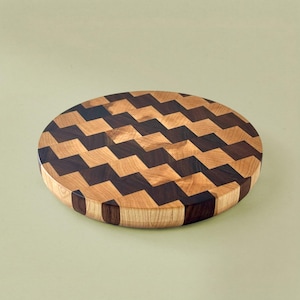 May include: Round wooden cutting board with a zigzag pattern. The board is made of alternating light and dark wood pieces, creating a visually appealing design. The board is approximately 12 inches in diameter.