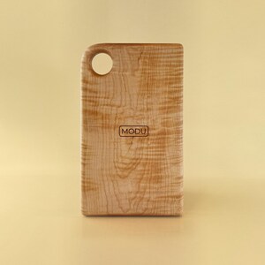 May include: A rectangular wooden cutting board with a rounded handle and a hole for hanging. The board has a light brown color with a natural wood grain pattern. The word "MODU" is printed on the front.