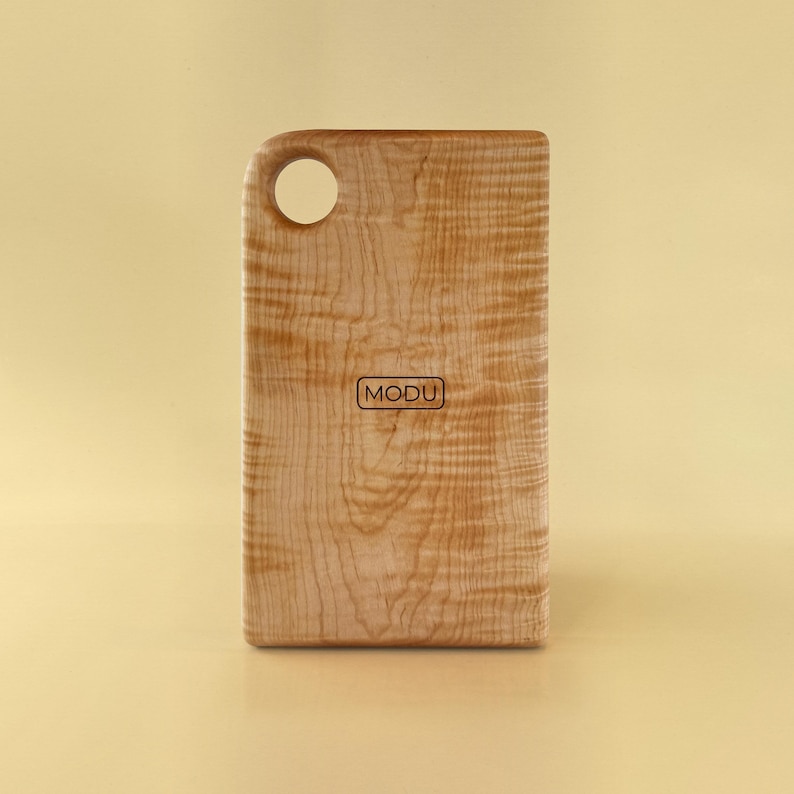May include: A rectangular wooden cutting board with a rounded handle and a hole for hanging. The board has a light brown color with a natural wood grain pattern. The word "MODU" is printed on the front.