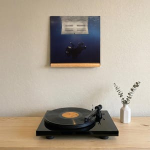 May include: A black turntable with a vinyl record playing, a wooden shelf holding a framed album cover with a diver in blue water, and a small white vase with greenery on a wooden table.