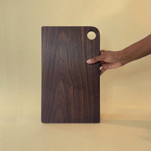 May include: A rectangular wooden cutting board with a dark brown finish and a rounded corner with a circular hole. The board is held by a hand against a light yellow background. The wood grain is visible.