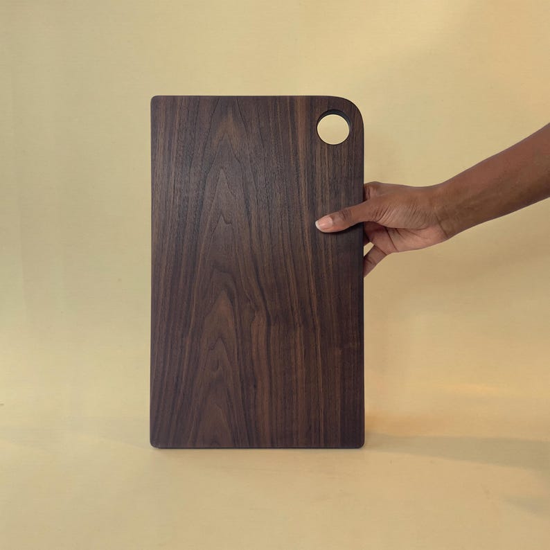 May include: A rectangular wooden cutting board with a dark brown finish and a rounded corner with a circular hole. The board is held by a hand against a light yellow background. The wood grain is visible.