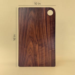 May include: A rectangular wooden cutting board with a dark brown finish. It measures 16 inches long and 10 inches wide, with a rounded corner and a circular hole for hanging. The wood grain is visible.