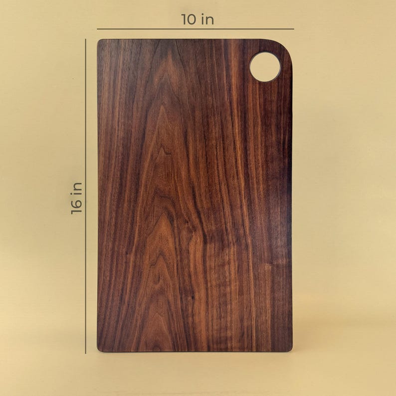 May include: A rectangular wooden cutting board with a dark brown finish. It measures 16 inches long and 10 inches wide, with a rounded corner and a circular hole for hanging. The wood grain is visible.