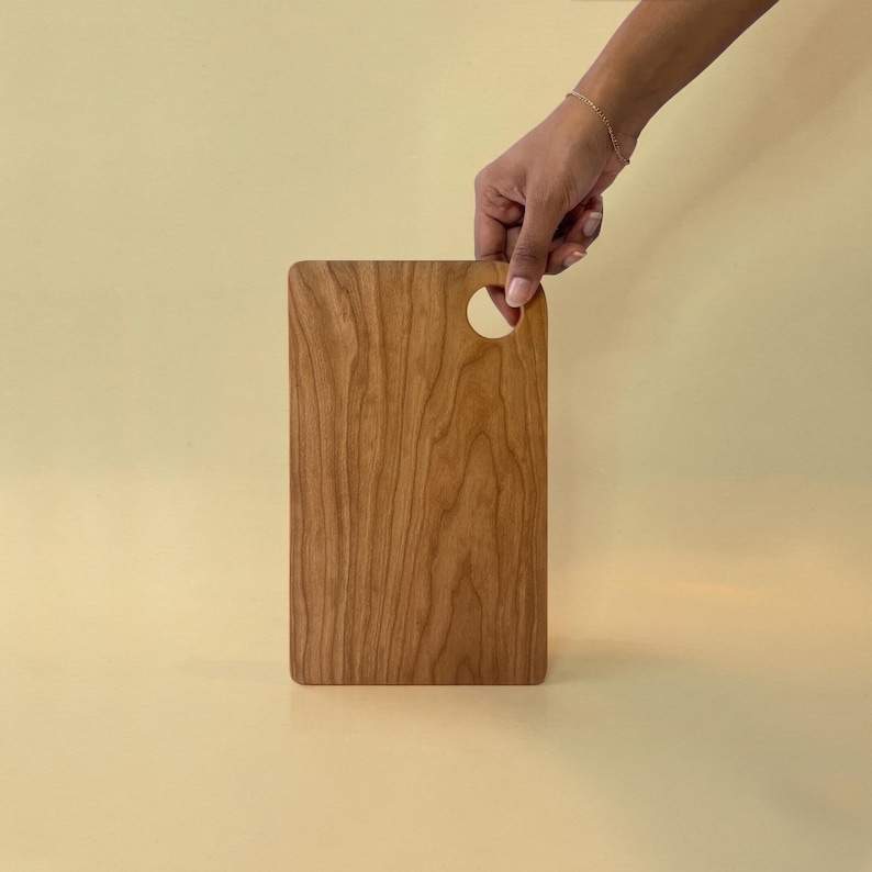 May include: A rectangular wooden cutting board with a rounded top and a circular handle. The board is made of light brown wood with a visible grain pattern. The board is being held by a hand.