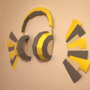 Puede incluir: Una obra de arte mural que presenta un par de auriculares grandes y estilizados. Los auriculares son principalmente grises y amarillos, con un diseño geométrico. Formas abstractas adicionales en gris, amarillo y beige están dispuestas cerca, creando una composición artística moderna.