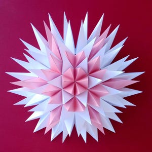 May include: A multi-layered paper star ornament featuring a geometric design with alternating white and light pink triangular points. The structure is composed of folded paper segments arranged in a radial pattern, creating a three-dimensional effect.