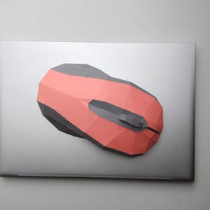 Könnte beinhalten: Eine rote und graue Computermaus liegt auf einem silbernen Laptop. Die Maus hat ein geometrisches Design mit roter Oberseite und grauen Seiten. Der Laptop ist geschlossen und hat eine glatte, metallische Oberfläche.