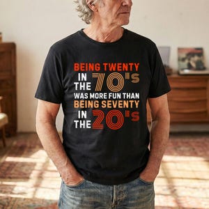 Könnte beinhalten: Schwarzes T-Shirt mit dem Text "BEING TWENTY IN THE 70'S WAS MORE FUN THAN BEING SEVENTY IN THE 20'S" in Orange, Braun und Weiß. Die Person trägt eine blaue Jeans. Der Hintergrund umfasst Holzmöbel und einen Vintage-Plattenspieler.