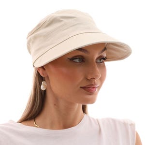 Könnte beinhalten: Beige Canvas-Cap mit Visor. Das Model trägt ein weißes ärmelloses Top und eine goldene Halskette. Die Ohrringe sind große, tropfenförmige Perlen. Der Hintergrund ist weiß.