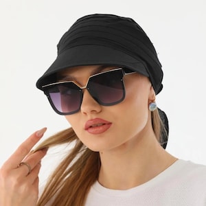 May include: Black sunglasses with a gold frame and gradient lenses. The sunglasses are worn with a black visor-style hat. The person is wearing a white shirt and a small earring.