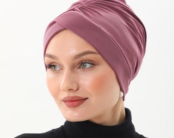 Puede incluir: Un turbante de color malva. La tela está fruncida en la parte superior y se envuelve alrededor de la cabeza. La persona de la imagen lleva un jersey de cuello alto negro. El fondo es blanco.
