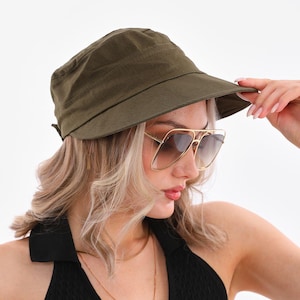 Peut inclure: Un chapeau de pêcheur vert olive à large bord. Le chapeau est porté avec des lunettes de soleil à monture dorée et un haut de style dos nu noir. Un collier en or est également visible. L'ensemble est posé sur un fond blanc.