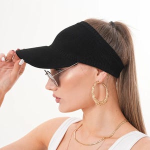May include: A black visor with a ribbed texture. The visor is worn with sunglasses, gold hoop earrings, and layered gold necklaces. The person is wearing a white vest top.