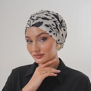 Może przedstawiać: Turban z kremowym tłem i czarnym wzorem kwiatowym. Ozdoba głowy jest noszona z czarną koszulą zapinaną na guziki i złotymi kolczykami. Na palcu widoczny jest pierścionek.