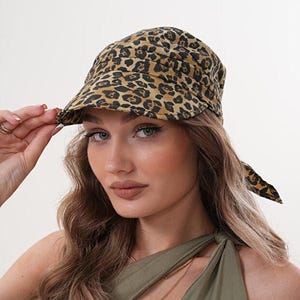Könnte beinhalten: Eine Kappe mit Leopardenmuster und gebogenem Schirm. Die Kappe ist beige und braun. Das Model trägt ein olivgrünes Oberteil und eine goldene Halskette. Die Kappe ist ein stilvolles Accessoire.
