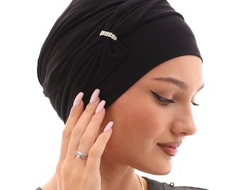 May include: Black turban-style head covering with a decorative rhinestone accent. The head covering is made of a soft, draped fabric. The person in the image is wearing a gray long-sleeve shirt and a gold necklace.