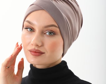 Puede incluir: Un turbante de color topo. La tela está fruncida en la parte superior, creando un efecto texturizado. La modelo lleva un cuello alto negro.