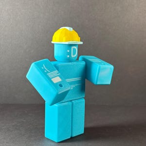 May include: A light blue, block-shaped figure with a yellow hard hat. The figure has a smiling face with the letter "D" and a white design on its left arm. The figure is standing with its arms out.