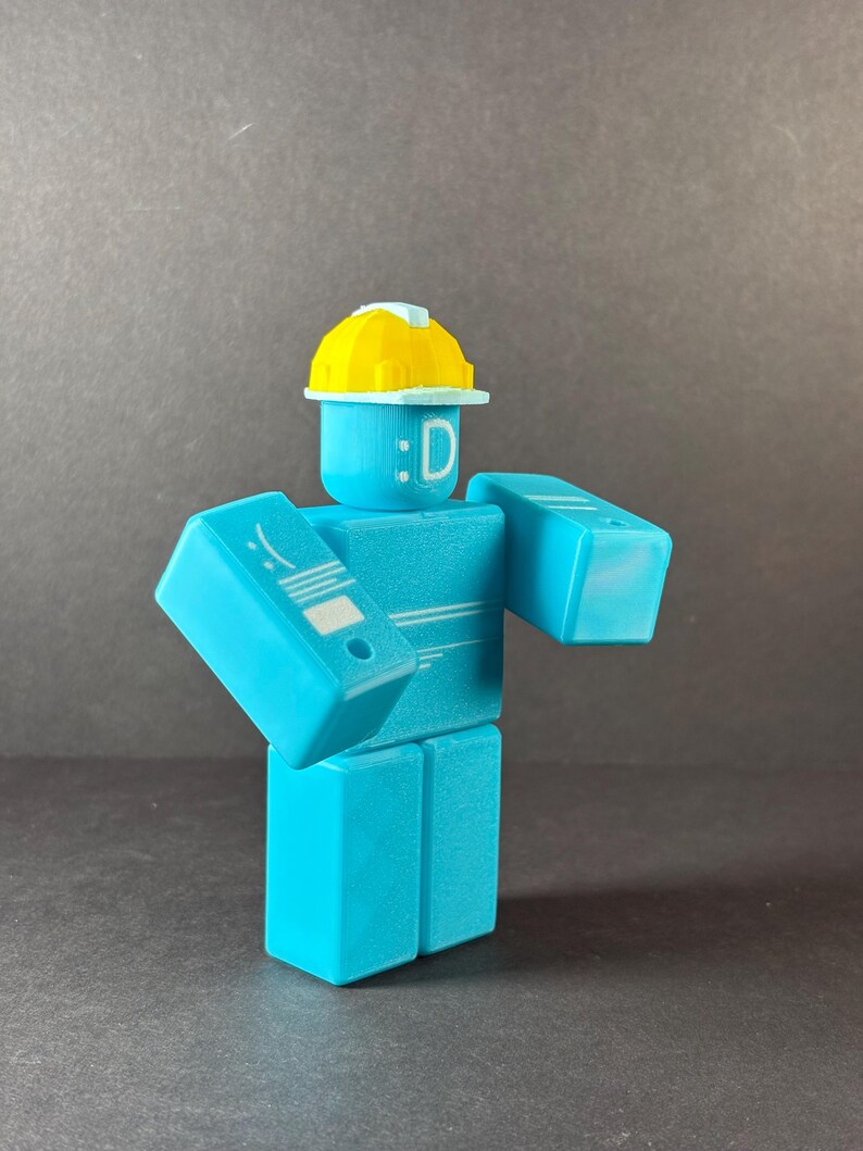 May include: A light blue, block-shaped figure with a yellow hard hat. The figure has a smiling face with the letter "D" and a white design on its left arm. The figure is standing with its arms out.