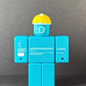 May include: A turquoise-colored block figure with a yellow hard hat. The head displays a smiling face with the letter "D". The figure has rectangular arms and legs, with a sad face on the left arm.