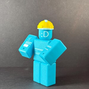 May include: A turquoise-colored, block-shaped figure with a yellow hard hat. The figure has a face with a colon and the letter "D". The figure's arms are angled outward. The figure is a 3D-printed toy.