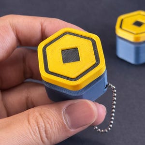 May include: A small, hexagonal keychain pendant with a yellow top and a black square in the center. The pendant has a blue base and a silver chain. Another similar pendant is visible in the background.