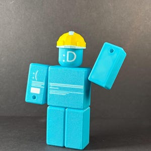 May include: A turquoise block figure with a yellow hard hat. The figure has a smiley face and the letter "D" on its head. The figure's arms are outstretched, and it has a sad face on one arm.