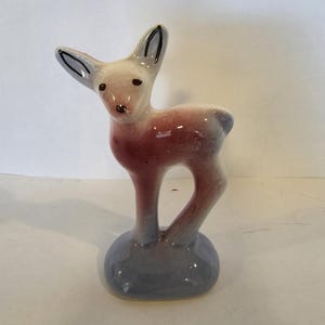 May include: A ceramic figurine of a deer, featuring a white head with black-lined ears and a reddish-brown body. The deer stands on a blue base. The figurine is a decorative item.
