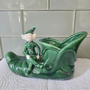 May include: A vintage green ceramic planter shaped like a shoe, with a seated elf figurine. The elf wears a green outfit and hat. The planter has a glossy finish and is likely for small plants or flowers.