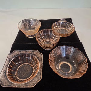May include: Five pieces of vintage pink depression glass tableware. The collection includes a small bowl with a floral design, a square dish with a floral border, and three other bowls with various ribbed and scalloped designs.
