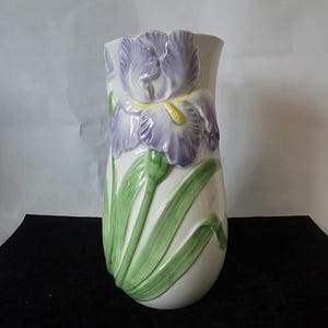May include: A white ceramic vase with a raised relief design of a purple iris flower and green leaves. The vase has a tall, slender shape and is suitable for displaying flowers. The iris has yellow accents.