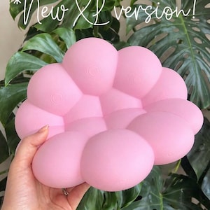 May include: A pink, cloud-shaped cushion with a textured surface. The cushion is held in a hand, with large green leaves in the background. Text in the image reads "New XL version!"